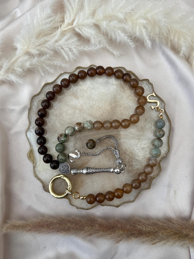 May include: A prayer bead strand featuring a mix of dark brown, tan, and mottled green stone beads. The strand includes a silver-toned metal tassel, a silver-toned letter S charm, and a gold-toned circular ring clasp at the end of the bead loop.