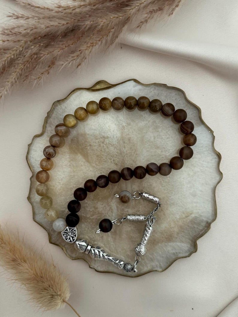 May include: A prayer bead strand featuring a gradient of smooth, round beads transitioning from light beige and golden yellow to deep dark brown. The strand includes silver-toned metal accents, a decorative shield-shaped charm, and a tassel component.