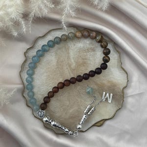 May include: A prayer bead strand featuring a gradient of smooth light blue, brown, and dark brown beads. The strand includes silver-toned metal accents, a decorative fleur-de-lis charm, a dangling silver letter M charm, and a textured silver tassel component.