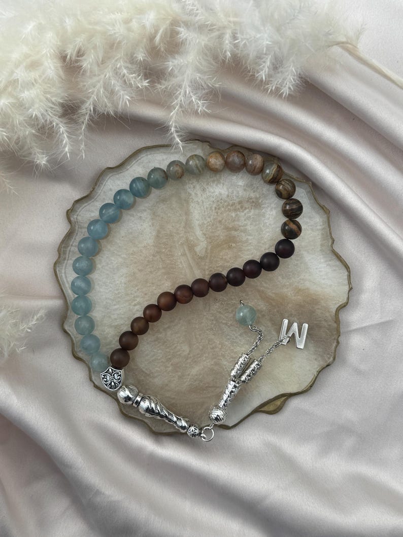 May include: A prayer bead strand featuring a gradient of smooth light blue, brown, and dark brown beads. The strand includes silver-toned metal accents, a decorative fleur-de-lis charm, a dangling silver letter M charm, and a textured silver tassel component.