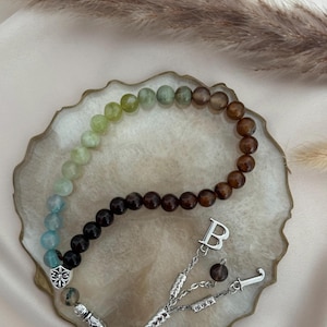 May include: A beaded prayer strand featuring a gradient of smooth, round stones transitioning from light blue and green to dark brown. The strand includes a silver-toned metal shield charm, a letter B charm, and a decorative silver-toned tassel end.