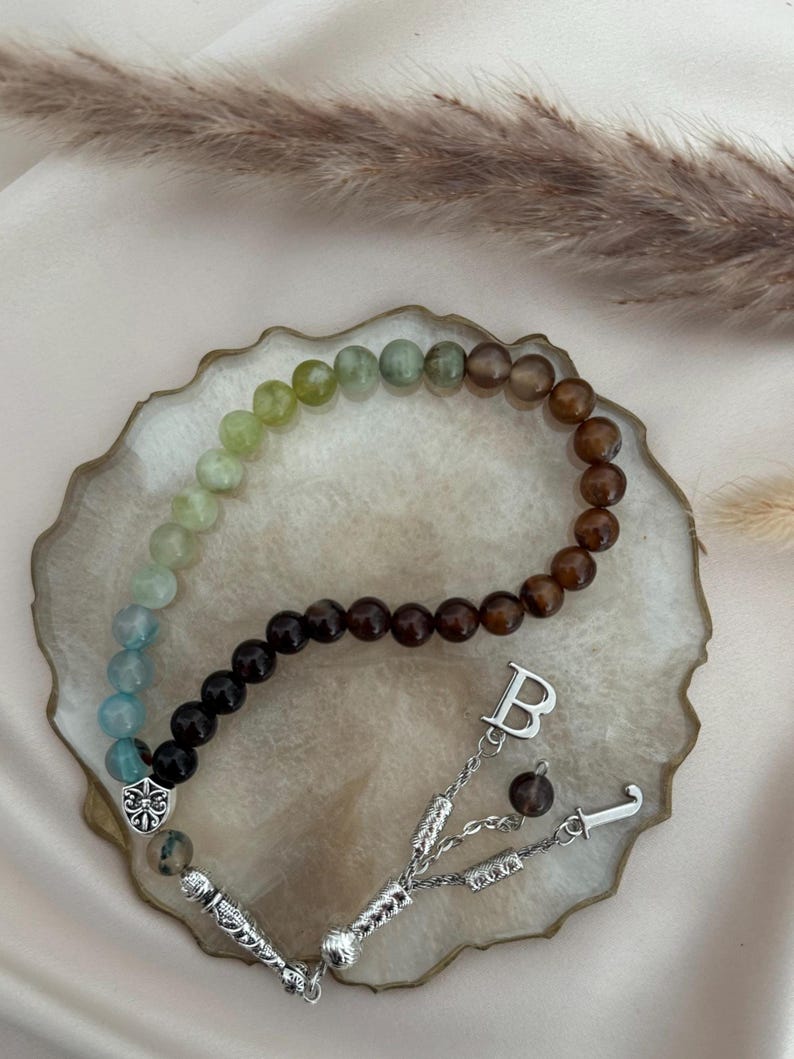 May include: A beaded prayer strand featuring a gradient of smooth, round stones transitioning from light blue and green to dark brown. The strand includes a silver-toned metal shield charm, a letter B charm, and a decorative silver-toned tassel end.