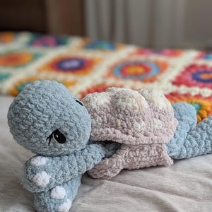 May include: A handmade crochet turtle plush toy in soft blue and pink hues. The turtle features a textured body, white spots, and black eyes. The plush is lying on a gray surface, with a colorful blanket in the background.