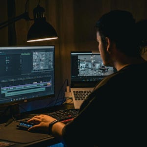 May include: A person working on a computer, editing video. The scene is dimly lit, with a large monitor displaying a video editing timeline and a smaller laptop screen showing a preview. A desk lamp provides focused light.