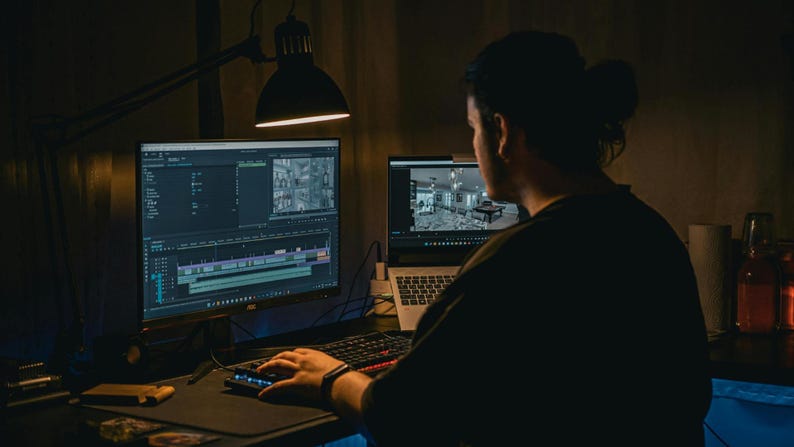 May include: A person working on a computer, editing video. The scene is dimly lit, with a large monitor displaying a video editing timeline and a smaller laptop screen showing a preview. A desk lamp provides focused light.