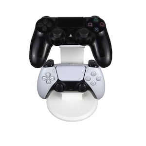 May include: A white vertical display stand holding two video game controllers. The top controller is black with a central touchpad and buttons, while the bottom controller is white with black accents, featuring a central touchpad and analog sticks.