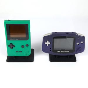 Op de afbeelding: Een groene Game Boy Color handheld console en een paarse Game Boy Advance handheld console, beide rechtop geplaatst op zwarte plastic displaystandaards. Elk apparaat is voorzien van een scherm, een richtingspad en knoppen aan de voorzijde.