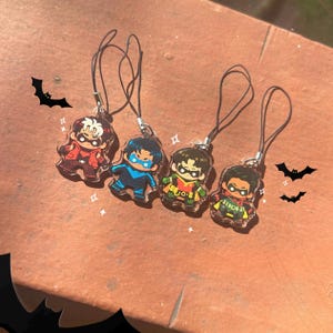 Könnte beinhalten: Vier Cartoon-Charms mit schwarzen Lanyards. Die Charms zeigen Charaktere in roten, blauen und grünen Kostümen mit schwarzen Masken. Dekorative Fledermaus-Silhouetten befinden sich im Hintergrund.