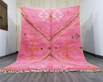 May include: A large, pink Moroccan rug with geometric designs. The rug features a central diamond pattern and cross-like shapes in orange and white, with zig-zag lines in orange and brown. The rug has a fringed edge.