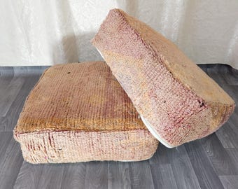 May include: Two square, textured floor cushions in shades of pink, yellow, and beige. One cushion is upright, the other is lying down. The cushions have a rectangular shape and are made of a woven material. The cushions are placed on a wooden floor.