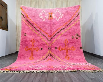May include: A large, pink Moroccan rug with a geometric design. The rug features orange, yellow, and brown accents, with cross-like and diamond-shaped patterns. The rug has a fringed edge and is displayed on a wooden floor.