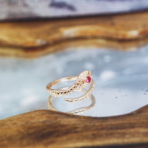 May include: A gold-coloured snake ring featuring a red gemstone eye. The ring is coiled, with detailed scale patterns. The ring is displayed on a reflective surface, showcasing its intricate design and craftsmanship.