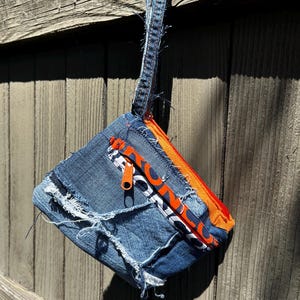 Puede incluir: Un estuche de mezclilla hecho a mano con un diseño de borde deshilachado. El estuche presenta una cremallera naranja y una correa de mezclilla. El tirador de la cremallera es naranja. El estuche está colgado en una valla de madera.