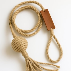 Puede incluir: Una correa de cuerda beige con un acento de cuero marrón. La correa presenta una bola anudada decorativa con una borla de hebras de cuerda sueltas. El diseño incluye un lazo y una pieza de cuero rectangular.