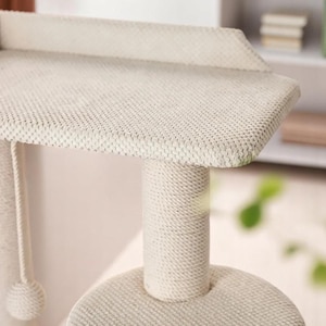 May include: A beige cat tree made with a textured, woven fabric. It has a rectangular top, a cylindrical support post, and a round base. A rope with a ball hangs from the side. The cat tree is designed for feline activity and relaxation.