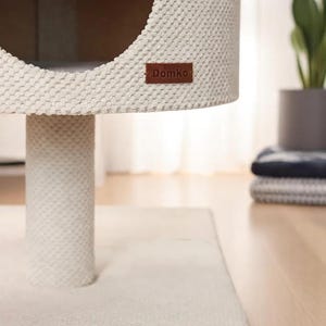 May include: A cream-coloured cat tree with a round opening and a textured surface. The cat tree has a cylindrical base and a rectangular base. A brown leather tag with the text "DomKo" is attached to the side. The cat tree is on a wooden floor.