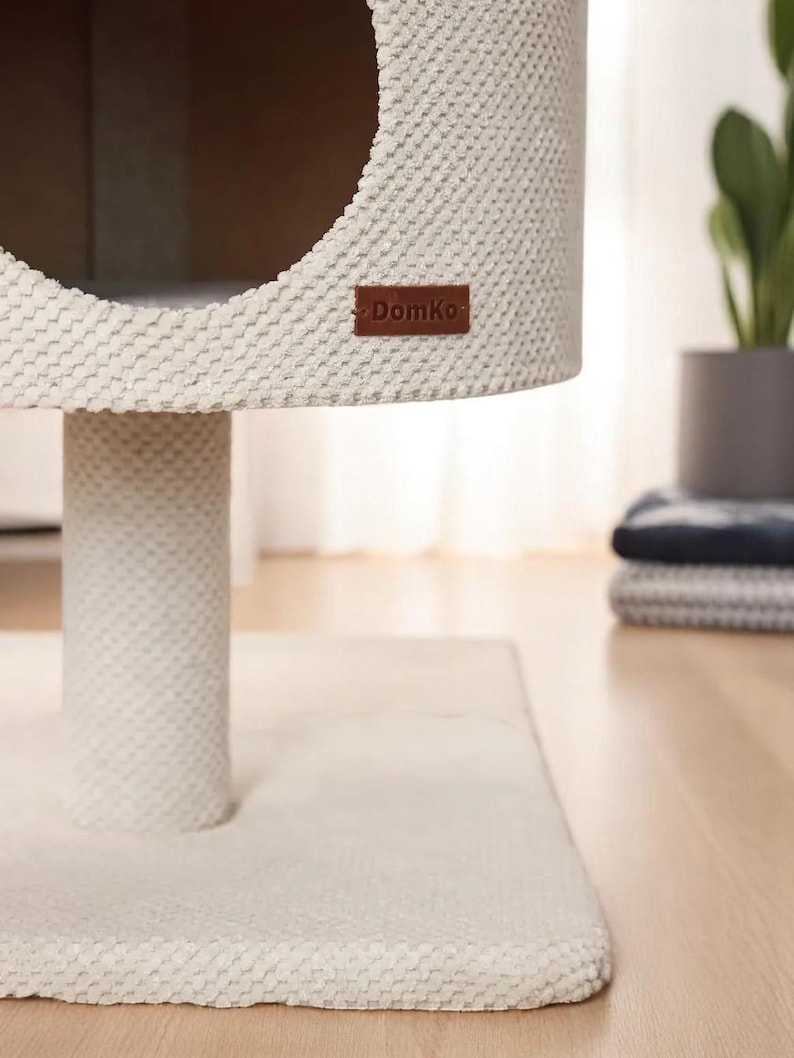 May include: A cream-coloured cat tree with a round opening and a textured surface. The cat tree has a cylindrical base and a rectangular base. A brown leather tag with the text "DomKo" is attached to the side. The cat tree is on a wooden floor.