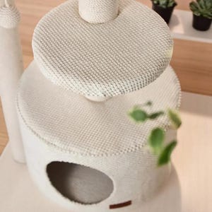 May include: A cream-coloured cat tree with a multi-tiered design. It features a circular base with an opening, a middle platform, and a top platform. The posts are wrapped in rope. The platforms are covered in a textured fabric.