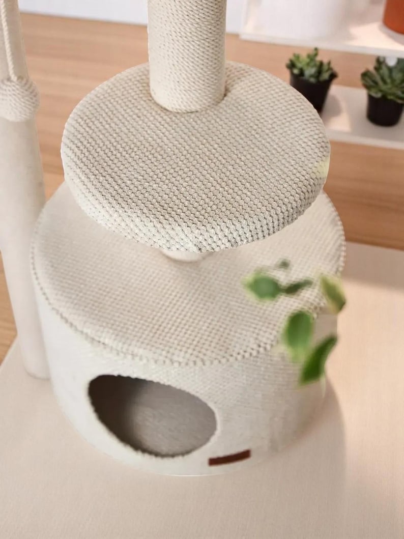 May include: A cream-coloured cat tree with a multi-tiered design. It features a circular base with an opening, a middle platform, and a top platform. The posts are wrapped in rope. The platforms are covered in a textured fabric.