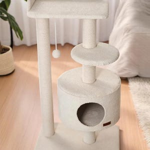 May include: A cream-coloured cat tree with a top platform, a mid-level perch, and a cylindrical hideaway with an opening. A dangling ball toy hangs from the top. The structure is made of textured fabric and supported by sturdy posts.