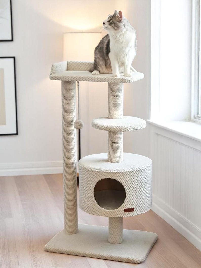 May include: A beige cat tree with multiple levels, including a top platform, a mid-level perch, and a base with a circular hideaway. A grey and white cat sits on the top platform. The posts are wrapped in sisal rope. The cat tree is designed for indoor cats.