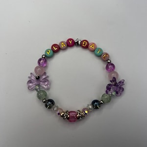 Puede incluir: Una pulsera de cuentas coloridas con la palabra "PLAYDATE" escrita en letras doradas. La pulsera presenta una variedad de cuentas en tonos rosa, morado, verde y transparente, con dijes decorativos de flores. Un accesorio divertido y alegre.