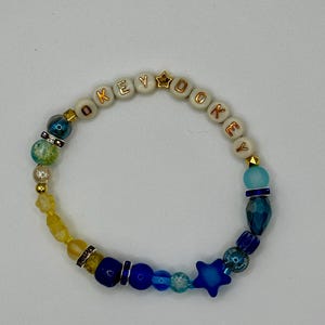 May include: A beaded bracelet with the word "OKEY DOKEY" spelled out in white letter beads. The bracelet features a variety of colorful beads in shades of blue, yellow, and gold, with a blue star charm. The bracelet is on a white background.