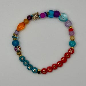 May include: A colorful beaded bracelet with the words "NUKA QUANTUM" spelled out in blue and red beads. The bracelet features a variety of beads in shades of orange, purple, blue, and gold, with a star-shaped charm.