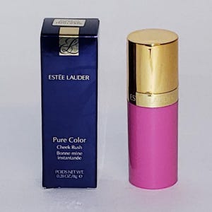 May include: A pink Estée Lauder Pure Color Cheek Rush blush stick with a gold cap, next to a dark blue box with the Estée Lauder logo and product information. The box reads "POIDS NET WT 0.28 OZ/8g e."