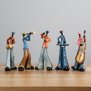 Può includere: Un set di cinque statuette decorative raffiguranti una band jazz. Ogni figura indossa abiti colorati e suona uno strumento diverso: sassofono, tromba, microfono, tastiera e contrabbasso. Le statuette sono poste su una superficie di legno.