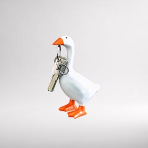 Peut inclure: Un porte-clés en forme d'oie blanche avec un bec et des pieds orange. Le porte-clés tient un jeu de clés sur un porte-clés argenté. Un article décoratif pour l'organisation de la maison.