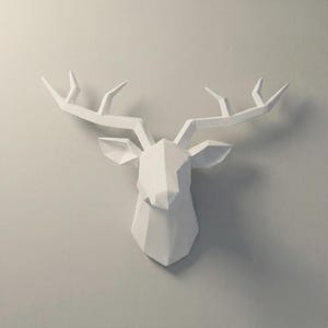 Könnte beinhalten: Ein weißer, geometrischer Hirschkopf als Wanddekoration. Die Skulptur hat ein polygonales Design mit detaillierten Geweihen und Ohren. Der Hirschkopf ist an einer hellgrauen Wand befestigt und schafft eine moderne und minimalistische Ästhetik. Ideal, um jedem Raum einen Hauch von naturnaher Kunst zu verleihen.