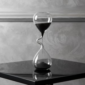 Puede incluir: Un reloj de arena de vidrio transparente con un centro curvo y arena negra. El reloj de arena está sobre una superficie negra y brillante. El fondo es una pared gris claro con un patrón sutil.