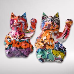 Könnte beinhalten: Zwei farbenfrohe Katzenskulpturen mit erhobenen Pfoten, die Graffiti-Kunst-Designs aufweisen. Die Skulpturen zeigen eine lebendige Mischung aus Farben, darunter Orange, Lila, Rosa und Blau, mit schwarzen Umrissen. Die Katzenfiguren haben spitze Ohren und stehen vor einem weißen Hintergrund.