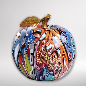 Puede incluir: Una escultura decorativa de manzana con un tallo y una hoja dorados. La manzana está cubierta de arte graffiti colorido, con tonos naranja, azul y rojo. La escultura tiene un acabado brillante y está sobre un fondo blanco.