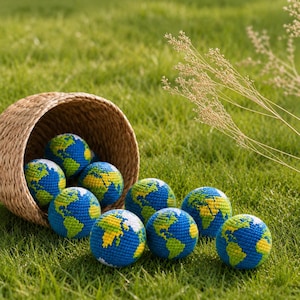 May include: A collection of crocheted globe-shaped balls in a woven basket, scattered on green grass. The balls feature blue oceans, green continents, and yellow landmasses. The basket is made of natural-colored woven material.