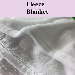 May include: Close-up of a white fleece blanket. The blanket has a soft, plush texture and is folded, showing the stitching along the edge. The words "Fleece Blanket" are printed in black at the top.