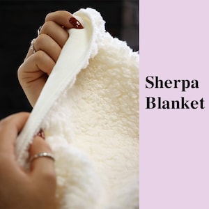 May include: Close-up of a cream-colored Sherpa blanket. The blanket has a soft, fluffy texture on one side and a smooth, satin-like material on the other. The text "Sherpa Blanket" is displayed in black.