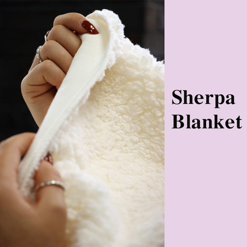 May include: Close-up of a cream-colored Sherpa blanket. The blanket has a soft, fluffy texture on one side and a smooth, satin-like material on the other. The text "Sherpa Blanket" is displayed in black.