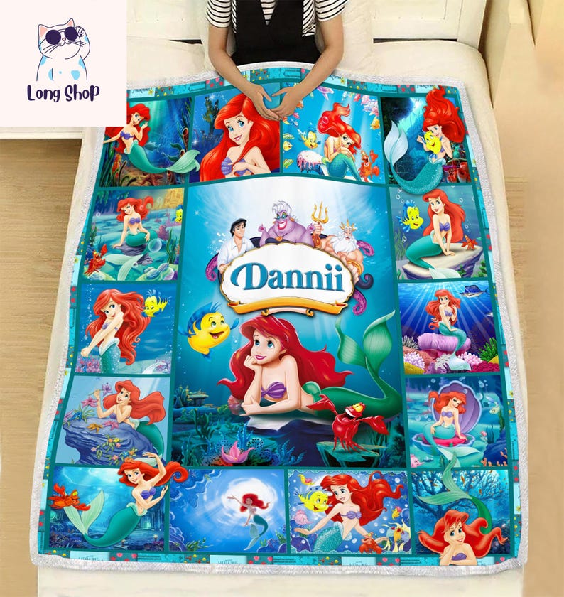May include: A turquoise and white blanket featuring multiple images of a mermaid with red hair, surrounded by sea creatures. The blanket has the name "Dannii" in a banner. The blanket is on a white bed.
