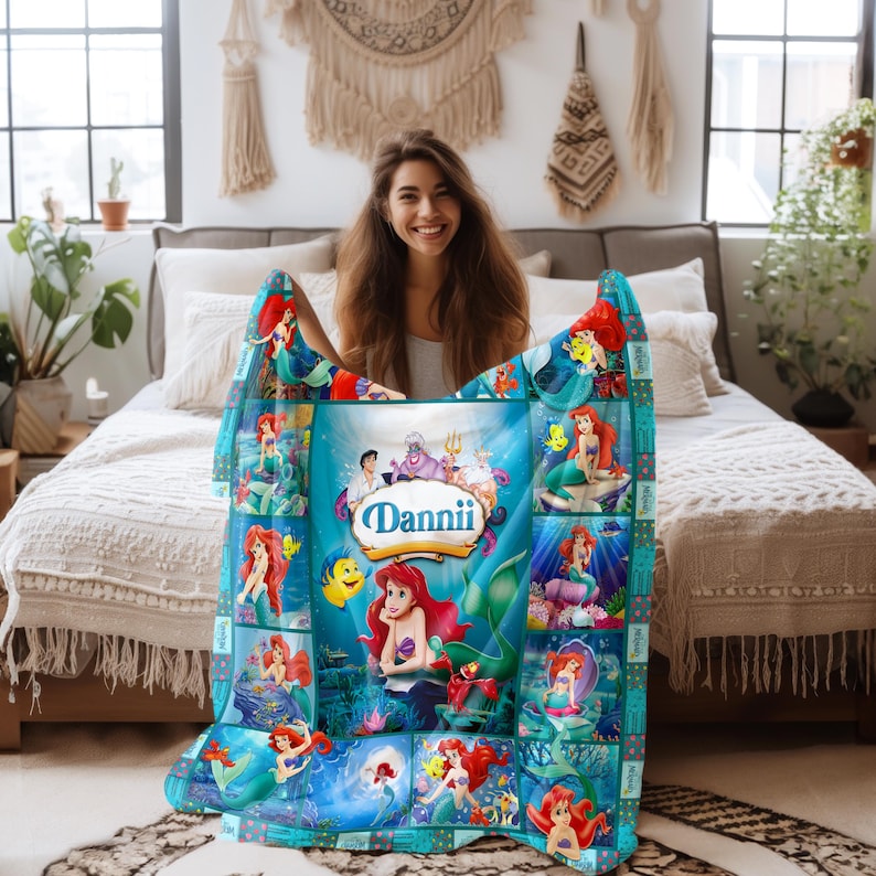 May include: A personalized, teal-colored blanket featuring various images of Ariel from The Little Mermaid. The blanket has a central panel with the name "Dannii" and other characters. The blanket is rectangular and appears to be made of soft material.