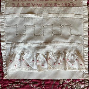 Peut inclure: Textile crème avec broderies. Le haut présente une bande avec l'alphabet et l'année 1923 en fil rouge. En dessous, des motifs géométriques et un bord à volants avec de petits points rouges.