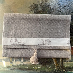 May include: A gray linen pouch with a white ribbon and a tassel. The ribbon has a sewing theme with images of scissors, thread, and a sewing machine. The text on the ribbon reads "A slow happy, handmade time in a modern world" and "My hobby is knitting and handicrafts".