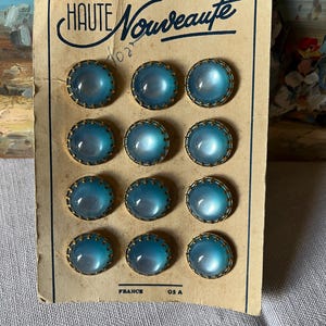 May include: A vintage card featuring twelve blue glass buttons with gold-coloured metal rims. The buttons are domed and have an opalescent quality. The card is labelled "HAUTE Nouveauté" and "FRANCE".