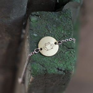 Puede incluir: Un brazalete dorado con un dije circular que presenta un dibujo simple de una rana. El brazalete está sobre una superficie de madera verde.