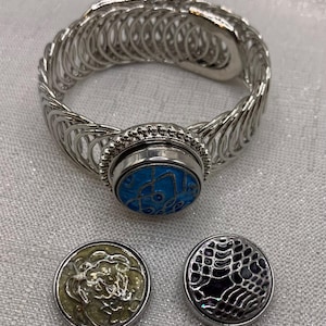 May include: A silver-toned bracelet with a large, round, blue and silver snap-on centerpiece. Two additional snap-on charms are included, one with a floral design and the other with a black and silver openwork pattern. The bracelet has a unique, open design.