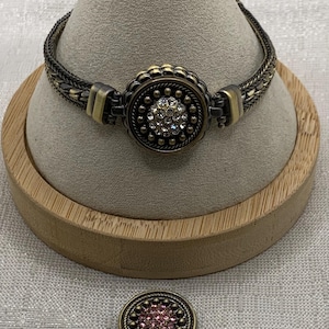 May include: An antique-style bracelet with a central round, ornate clasp. The clasp features a cluster of clear crystals surrounded by a decorative border. The bracelet has a woven chain design in a bronze and gold color. A matching decorative button is also present.