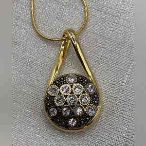 May include: Gold-toned pendant necklace with a teardrop design. The pendant features a circular element with a black background, adorned with numerous clear, sparkling crystals. The necklace chain is a delicate gold color.