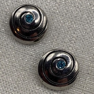 May include: Two circular silver-toned metal buttons featuring a raised spiral design that leads to a small, bright blue rhinestone set in the center of each piece. The buttons have a polished, reflective finish and a smooth, rounded edge.
