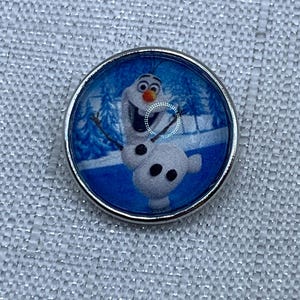 May include: A circular silver-toned button featuring a glossy graphic of a white snowman character with an orange carrot nose and black coal buttons, set against a blue winter background with stylized trees and snow-covered ground.